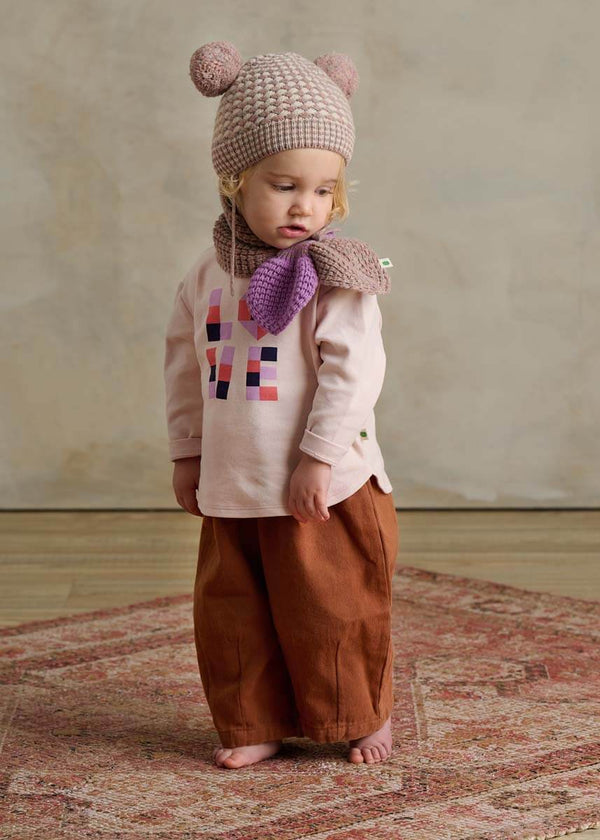 Child wearing a pink knit hat with bear ears, brown pants, and a colorful shirt 