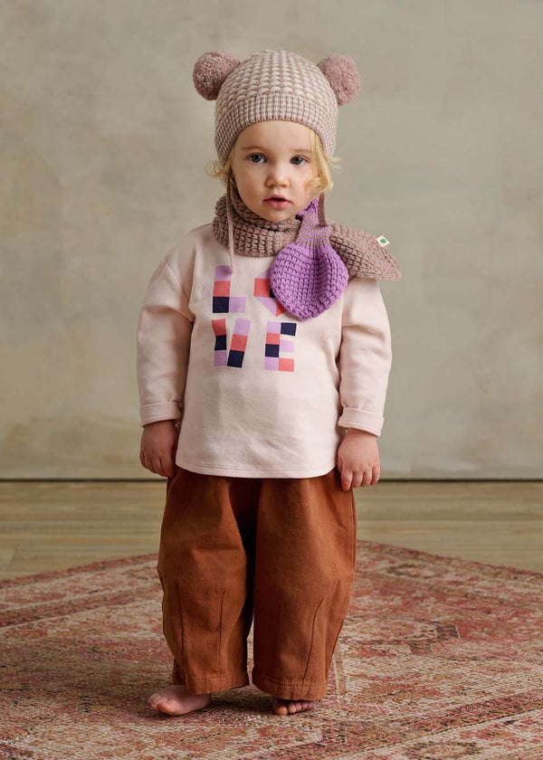 Child wearing a pink knit hat with bear ears, pink cotton long-sleeve t-shirt with colorful design, brown pants, and purple scarf with heart.