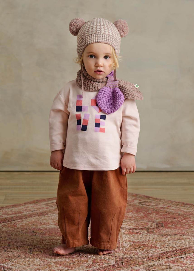 Child wearing a pink knit hat with bear ears, pink cotton long-sleeve t-shirt with colorful design, brown pants, and purple scarf with heart.