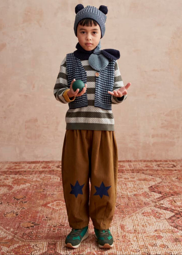 Child wearing a striped sweater, brown pants with star patterns, and a knitted hat with pom-poms 